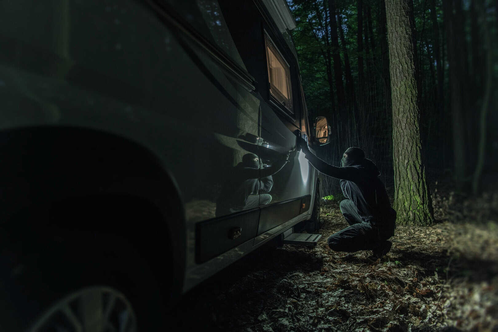 Man Fully Dressed in Black With Covered Face Trying to Break Into a Camper Van Parked in the Woods at Night. Robbery During Traveling. Criminal Theme.