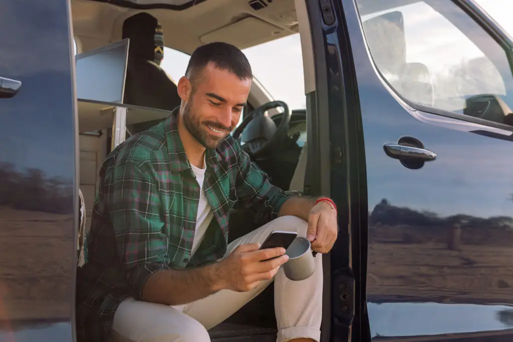 smiling young man using his mobile phone sitting on the door of his camper van, concept of freedom and digital nomad lifestyle