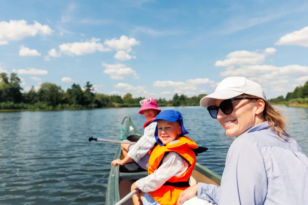 Woman two children enjoy having fun paddling canoe rent across lake river water wearing bright orange life vests hot sunny summer day. Healthy recreational activity lifestyle vacation trip journey.